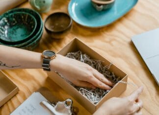 Zdrowe prezenty dla pracowników – pakiety well-being, sport i relaks Kobieta pakuje kartonowe pudełko z wypełnieniem na drewnianym biurku