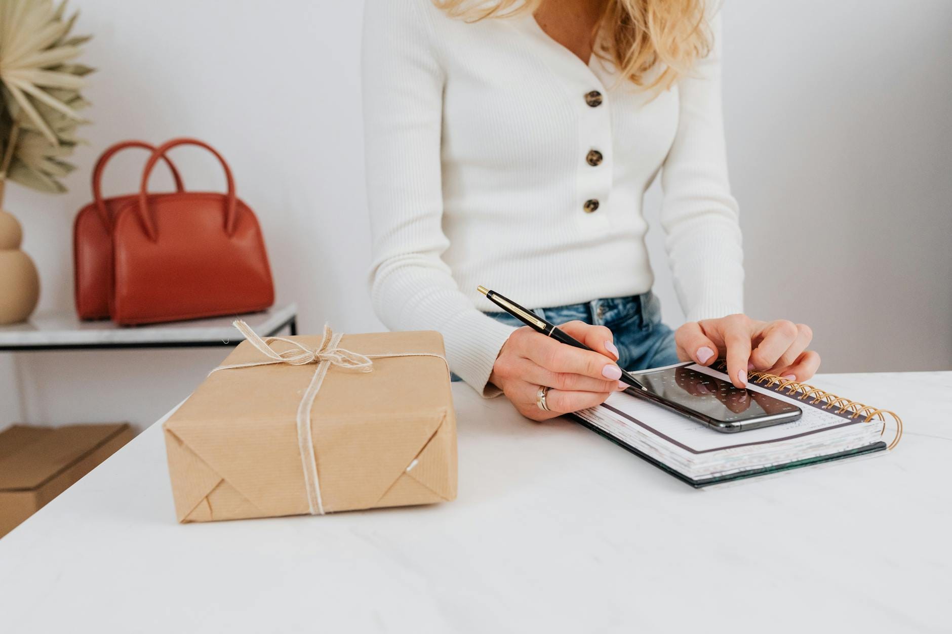 Kobieta planuje prezenty firmowe przy smartfonie, notesie i pudełku z prezentem